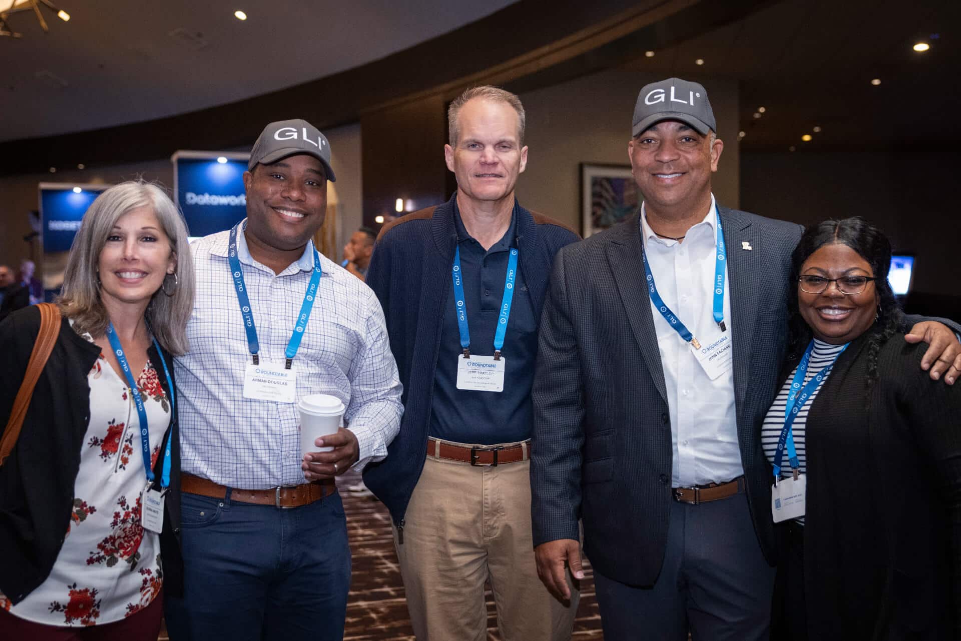Five conference attendees, wearing professional attire, as well as a GLI-branded hat, pose for a photo in the entrance of the Regulators Roundtable.