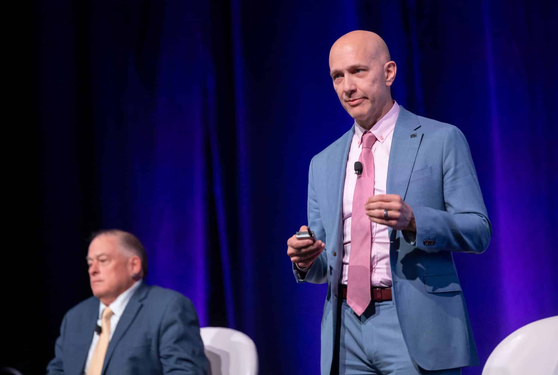 Gus Fritschie, wearing a blue suit, delivers a presentation on stage, holding a remote, with panelists seated behind him.