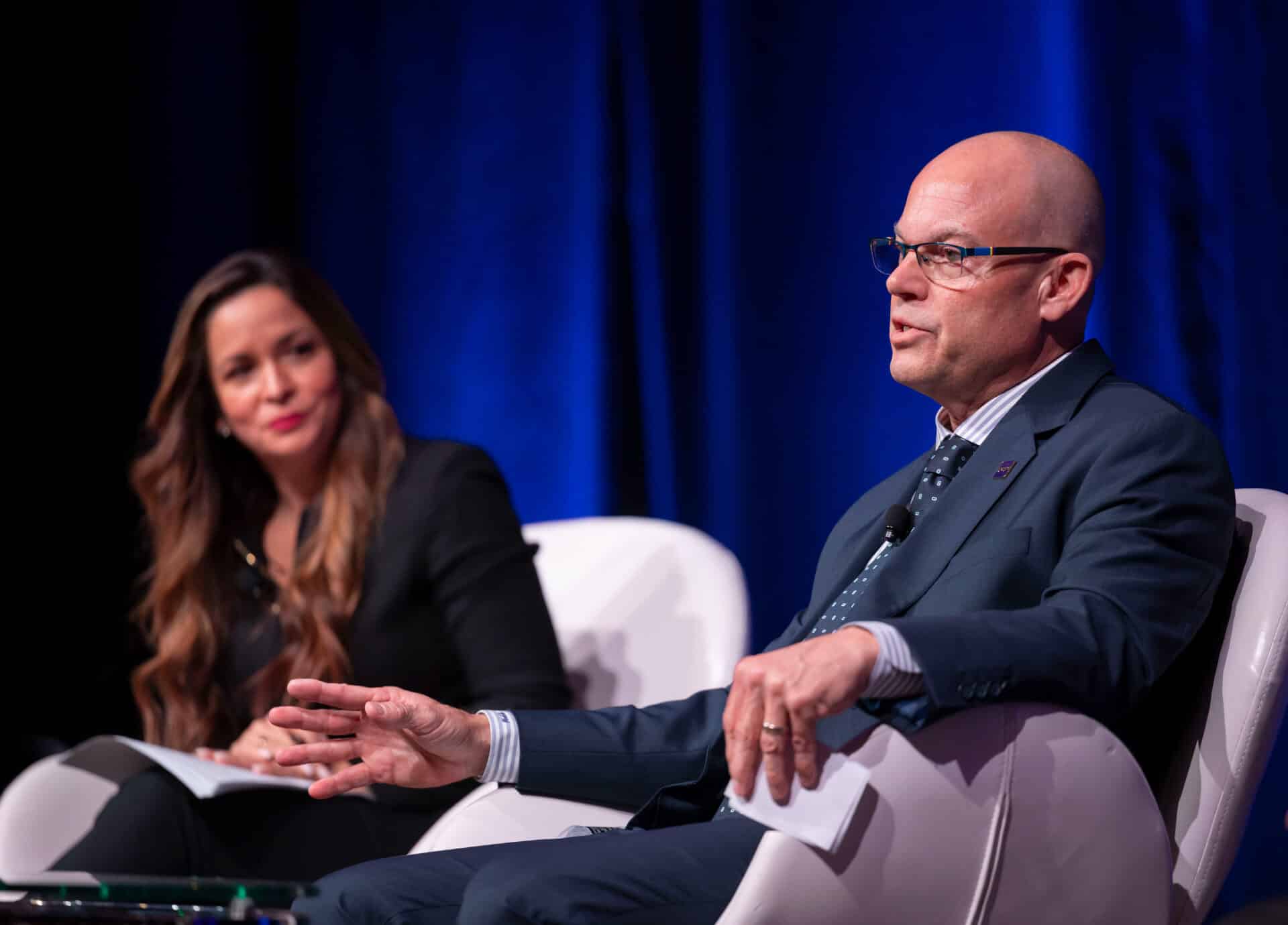 Two conference panelists seated on stage during a professional discussion, speaking into microphones against a blue-lit backdrop.