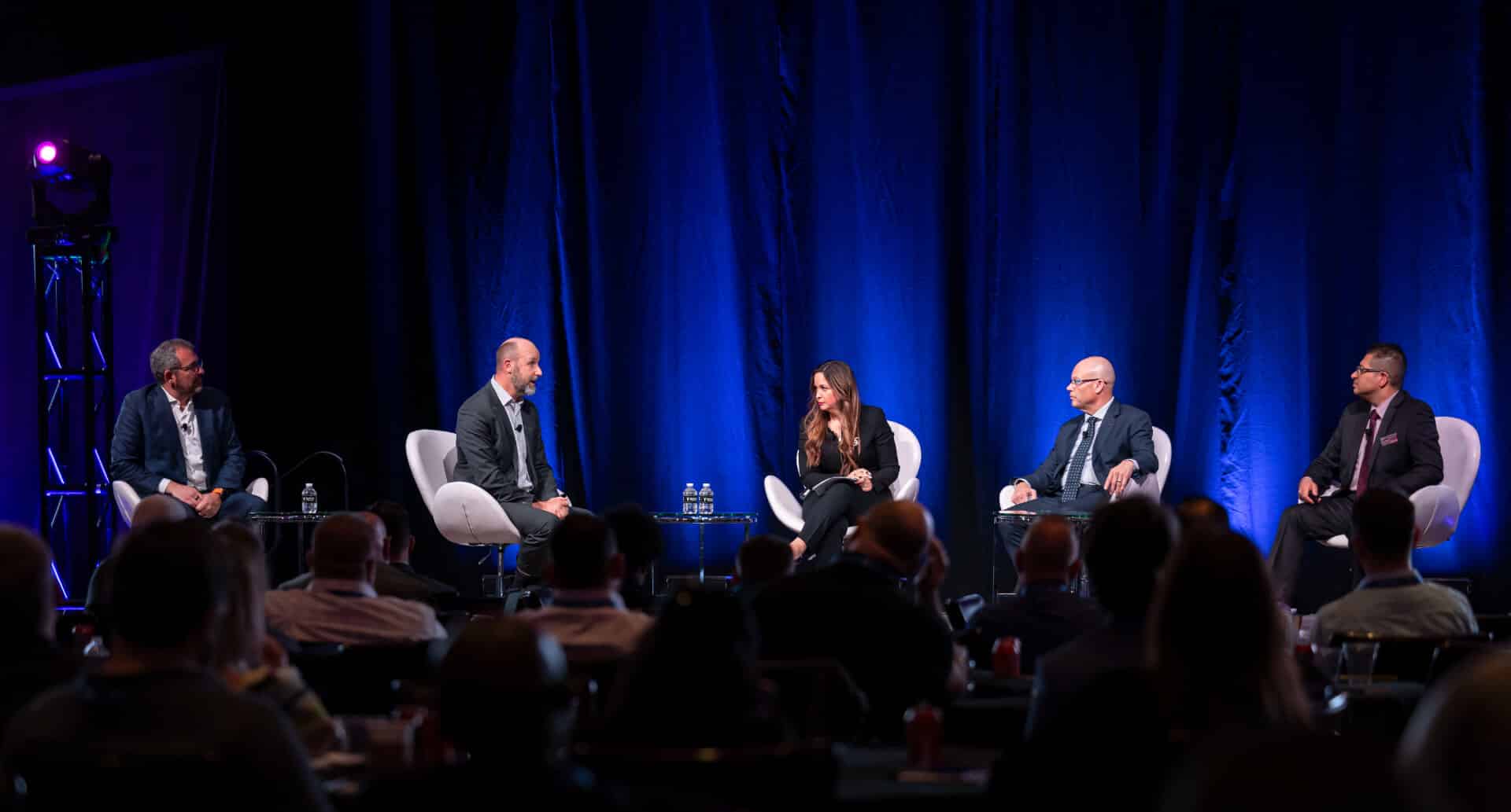 Multiple panelists, including Karen Marcela Sierra-Hughes participate in a formal conference session on stage during the Regulators Roundtable, seated in white chairs under professional lighting.