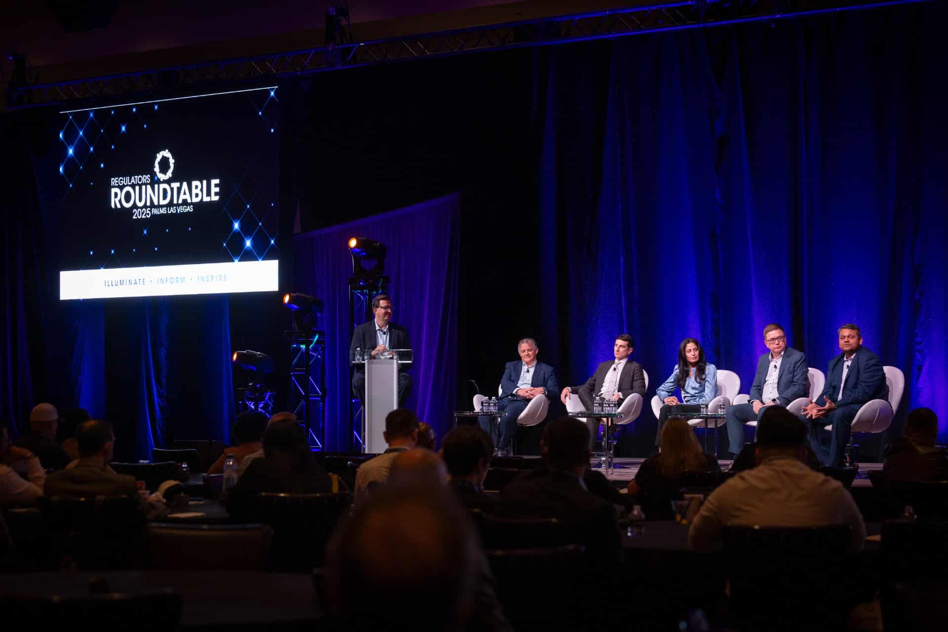 A wide view of five panelists and a moderator on stage during a discussion at the Regulators Roundtable, with a large screen and blue lighting behind them.
