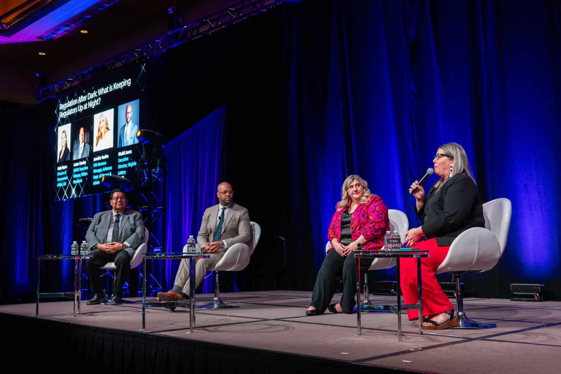 Multiple panelists participate in a formal conference session on stage during the Regulators Roundtable, seated in white chairs under professional lighting.