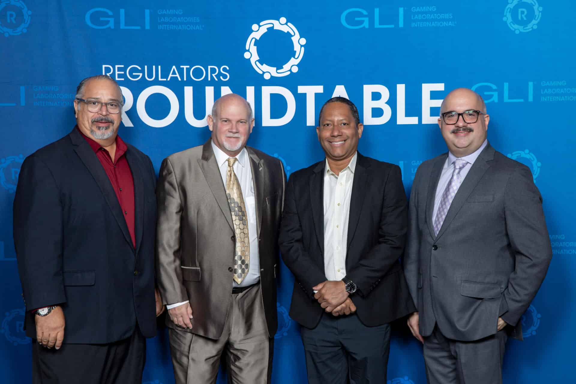 A group of four gaming professionals pose together for a photo in front of a blue Regulators Roundtable backdrop with GLI logos interpolated throughout.