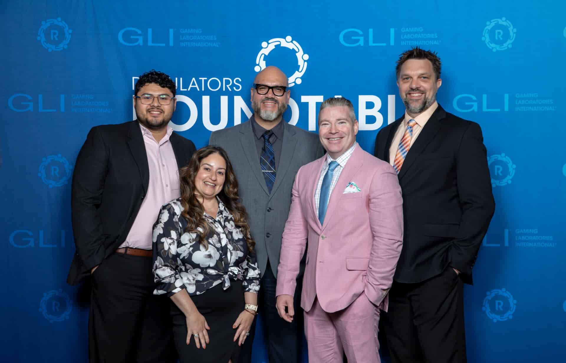 A group of five gaming professionals pose together for a photo in front of a blue Regulators Roundtable backdrop with GLI logos interpolated throughout.