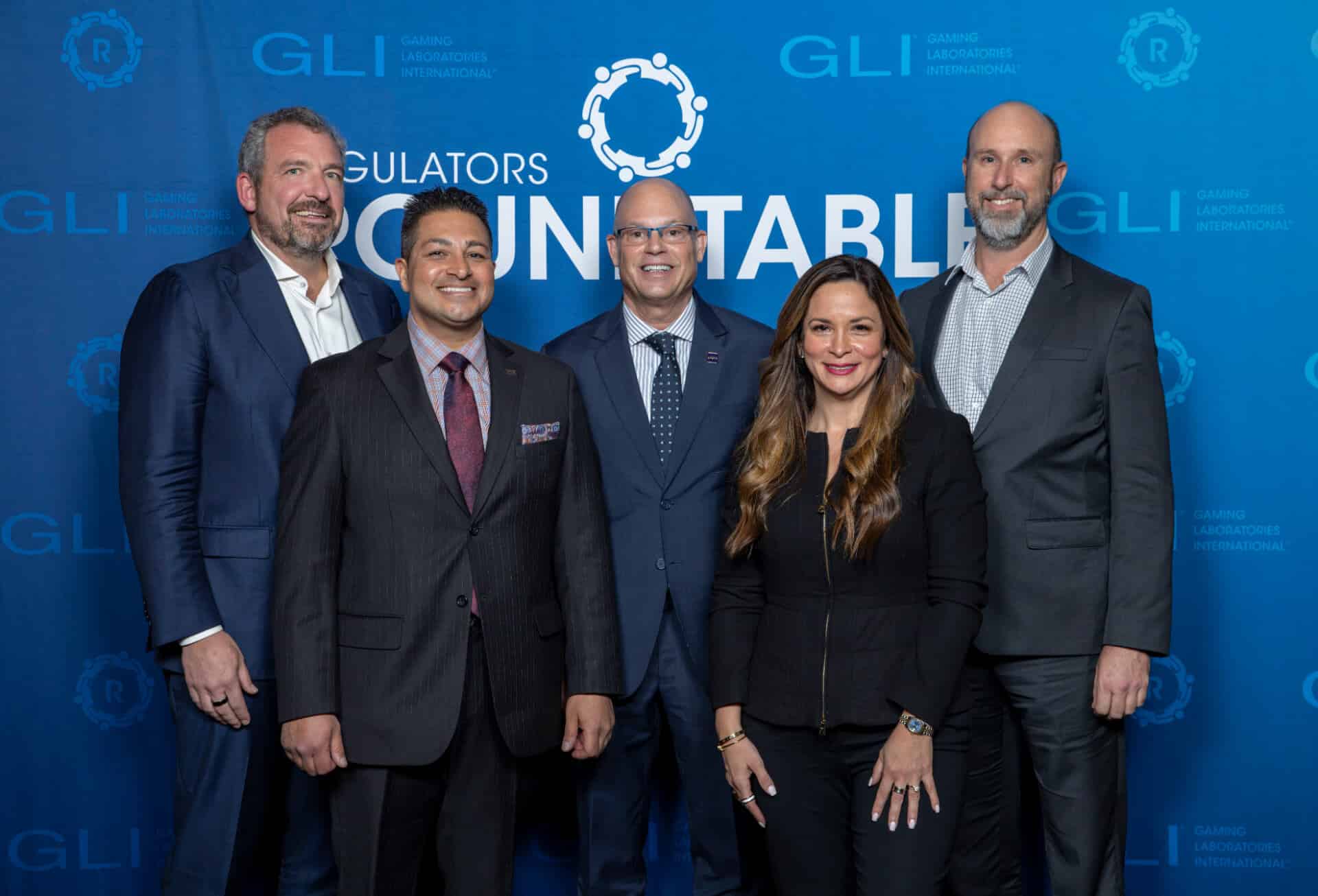 A group of five gaming professionals pose together for a photo in front of a blue Regulators Roundtable backdrop with GLI logos interpolated throughout.