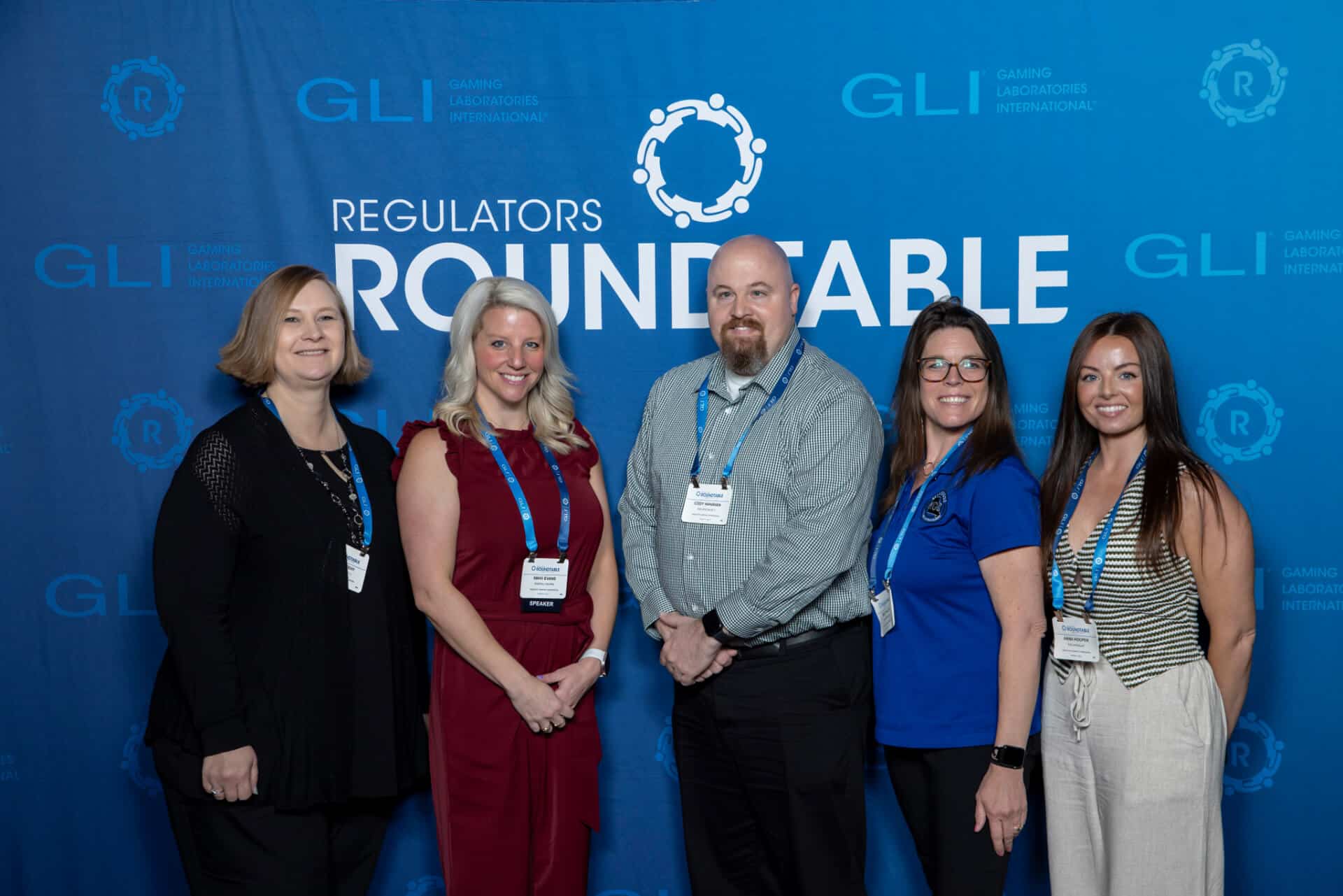 A group of five gaming professionals pose together for a photo in front of a blue Regulators Roundtable backdrop with GLI logos interpolated throughout.