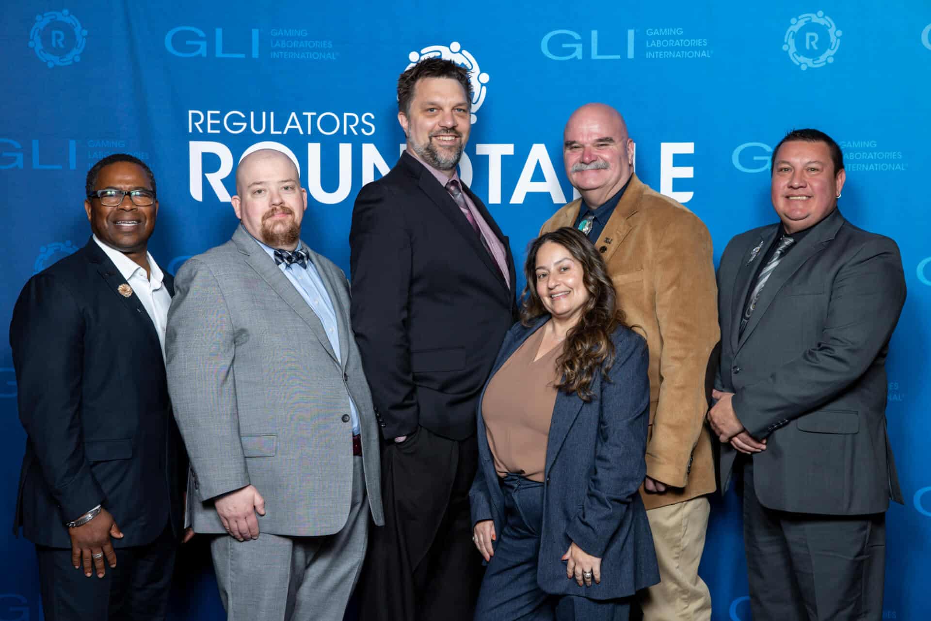 A group of six gaming professionals pose together for a photo in front of a blue Regulators Roundtable backdrop with GLI logos interpolated throughout.