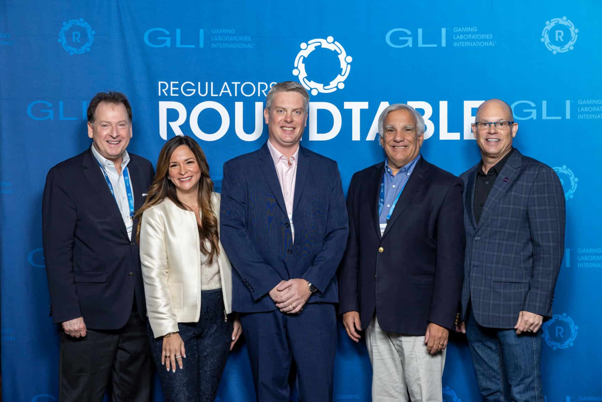 A group of five gaming professionals pose together for a photo in front of a blue Regulators Roundtable backdrop with GLI logos interpolated throughout.