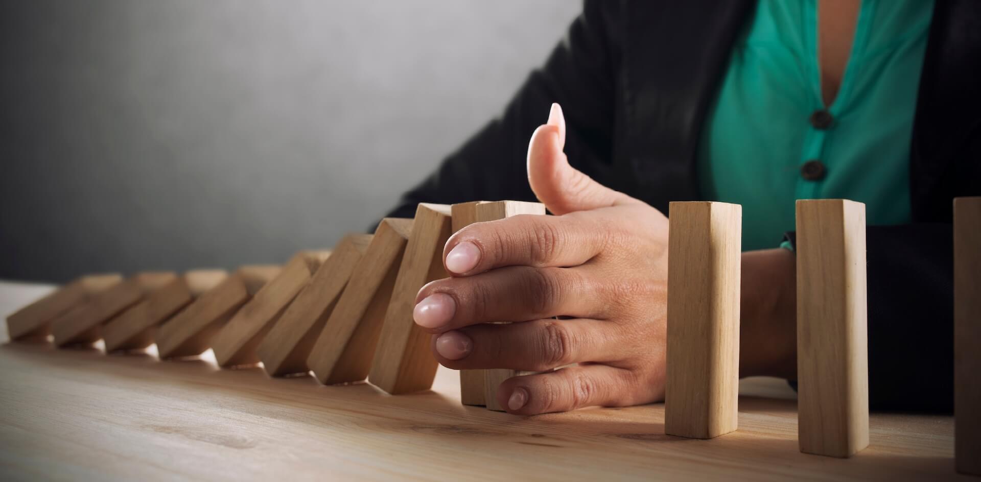 A person's hand stops a line of falling wooden dominoes on a table, symbolizing intervention and control to prevent negative outcomes in the context of responsible gaming.